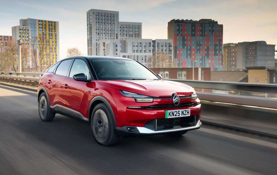 Red Citroën ë-C4 driving on an urban road with modern buildings in the background under a clear sky.