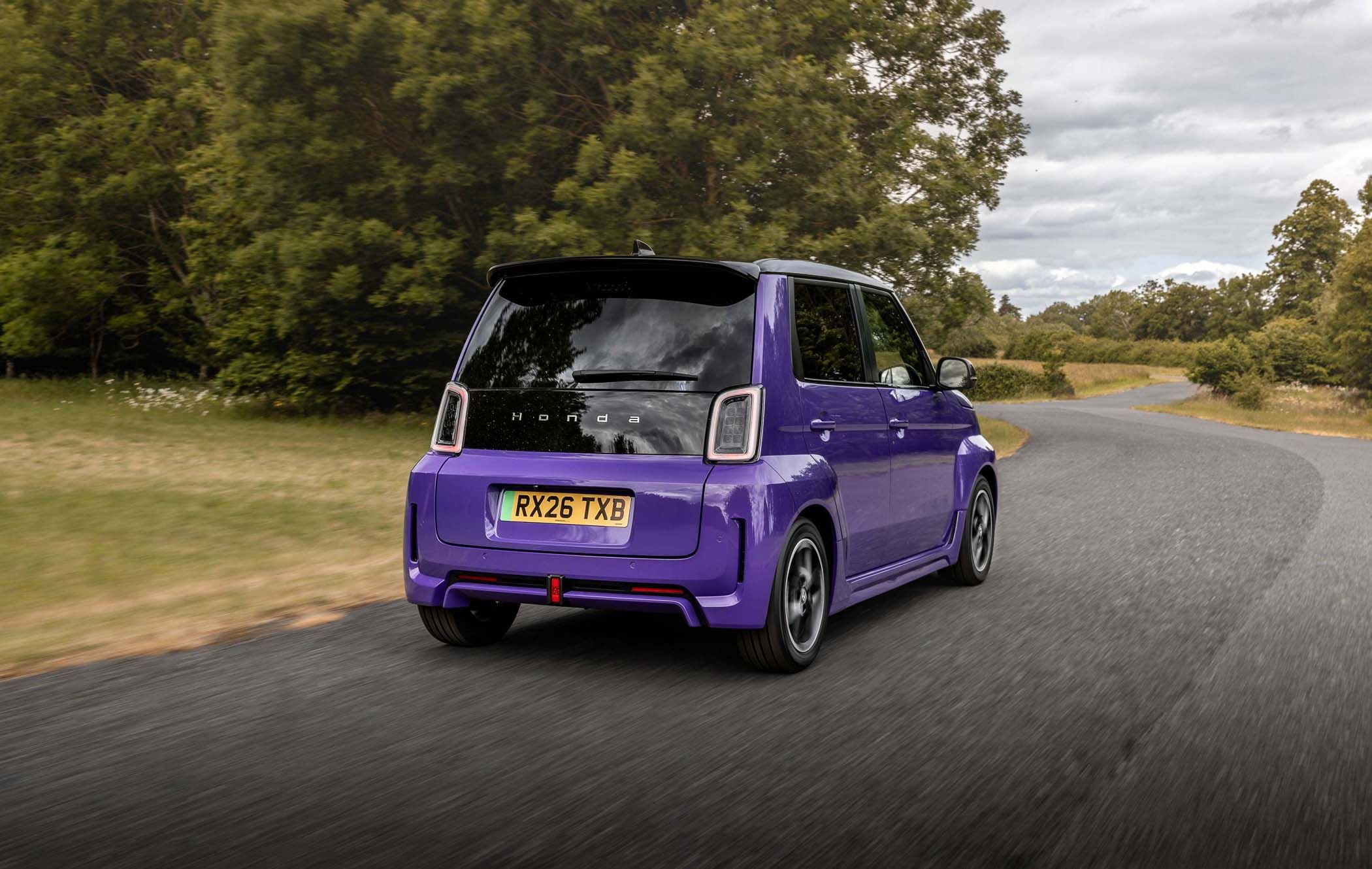 A purple Honda Super-N drives on a curved country road, surrounded by trees and grass under a cloudy sky.