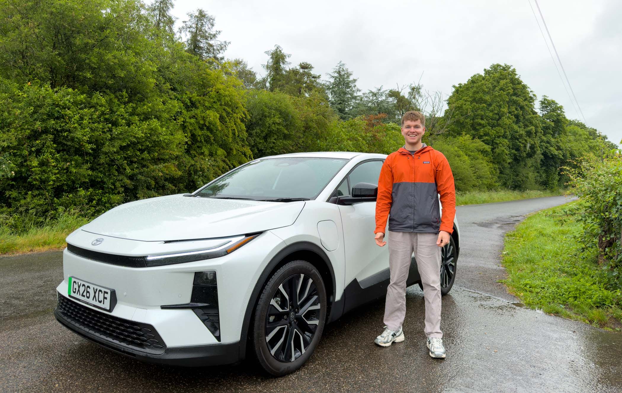 Craig Hale stands beside a white car on a rural road, surrounded by lush green trees under a cloudy sky.