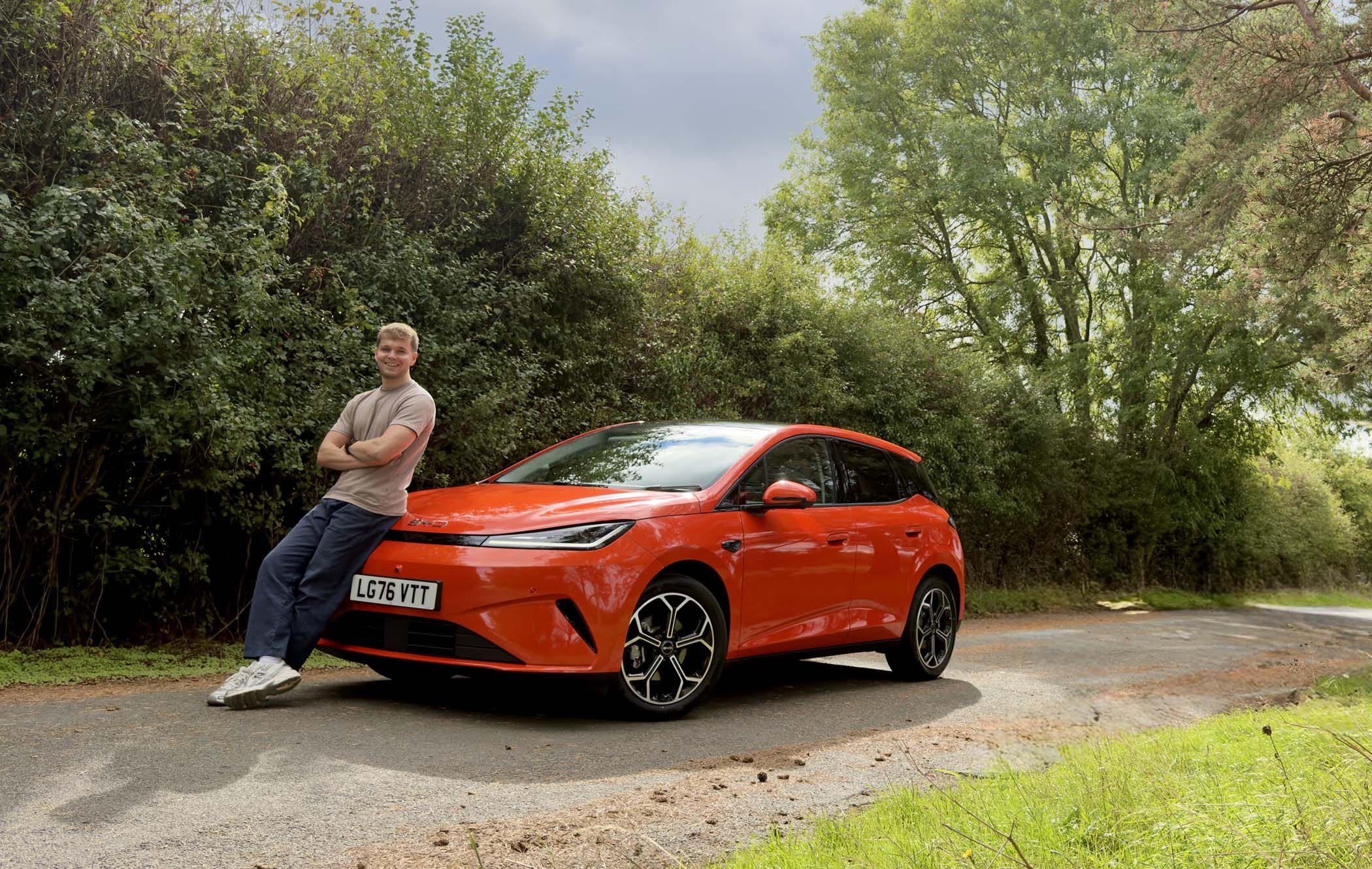 Craig Hale leaning on an orange BYD DOLPHIN G DM-i parked on a rural road, surrounded by green trees and bushes.