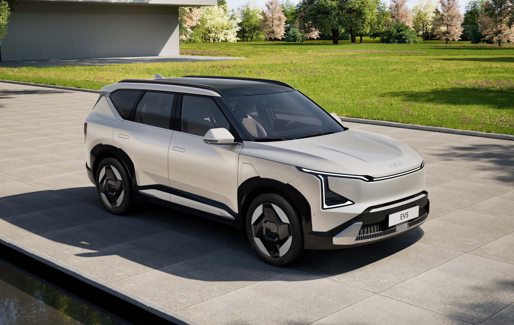 Silver Kia EV5 parked on a paved area, surrounded by green grass and trees under a clear sky.