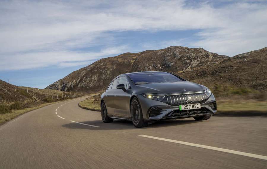 A grey Mercedes-AMG EQS 53+ drives along a winding road through a mountainous landscape under a partly cloudy sky.