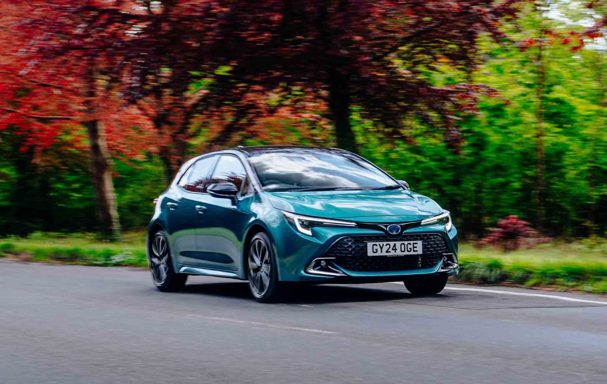 A teal Toyota Corolla driving on a road lined with trees, featuring green and red foliage, under a clear sky.