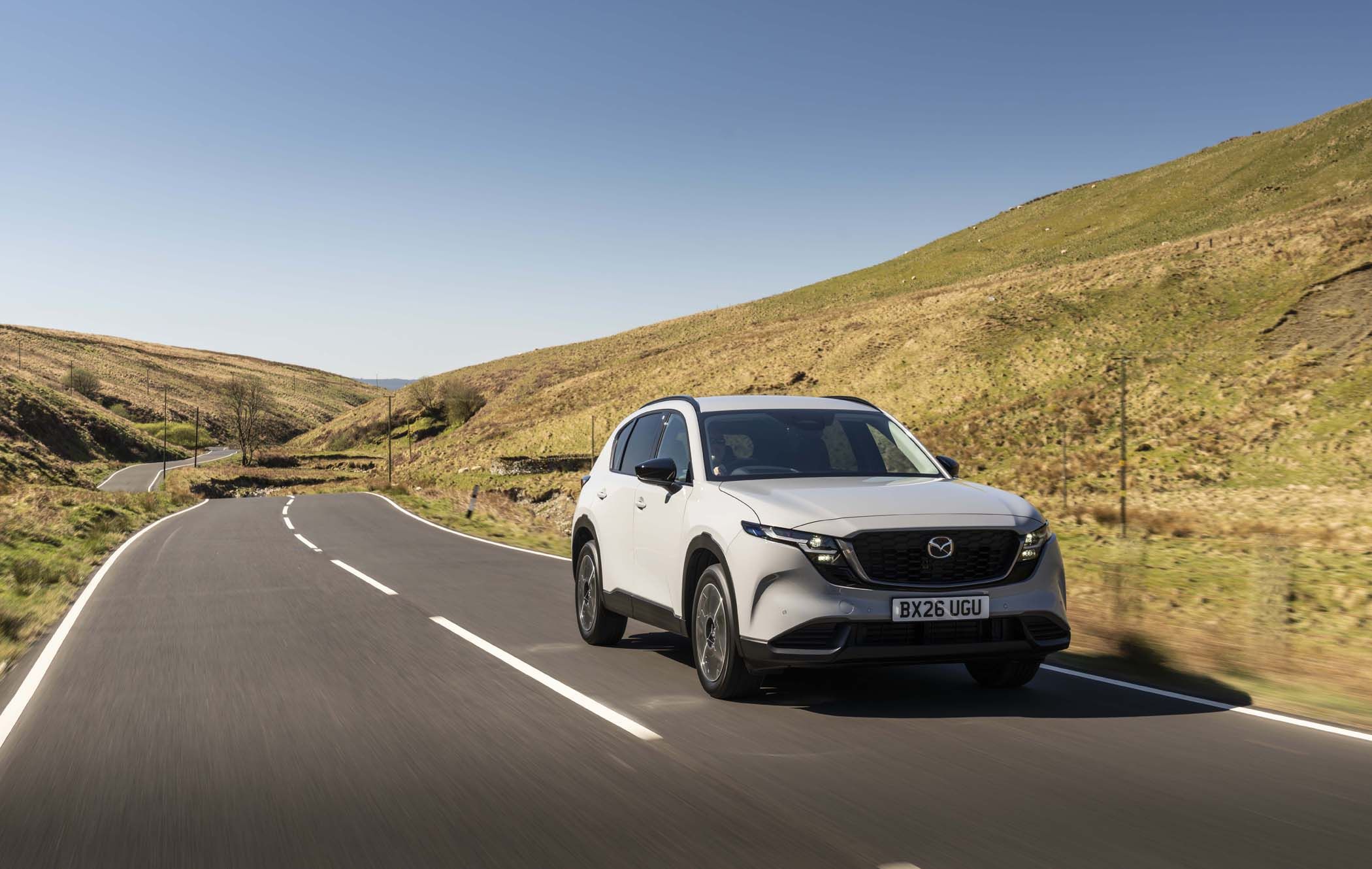 Grey Mazda CX-5 driving on a curvy road through a hilly, grassy landscape under a clear blue sky.