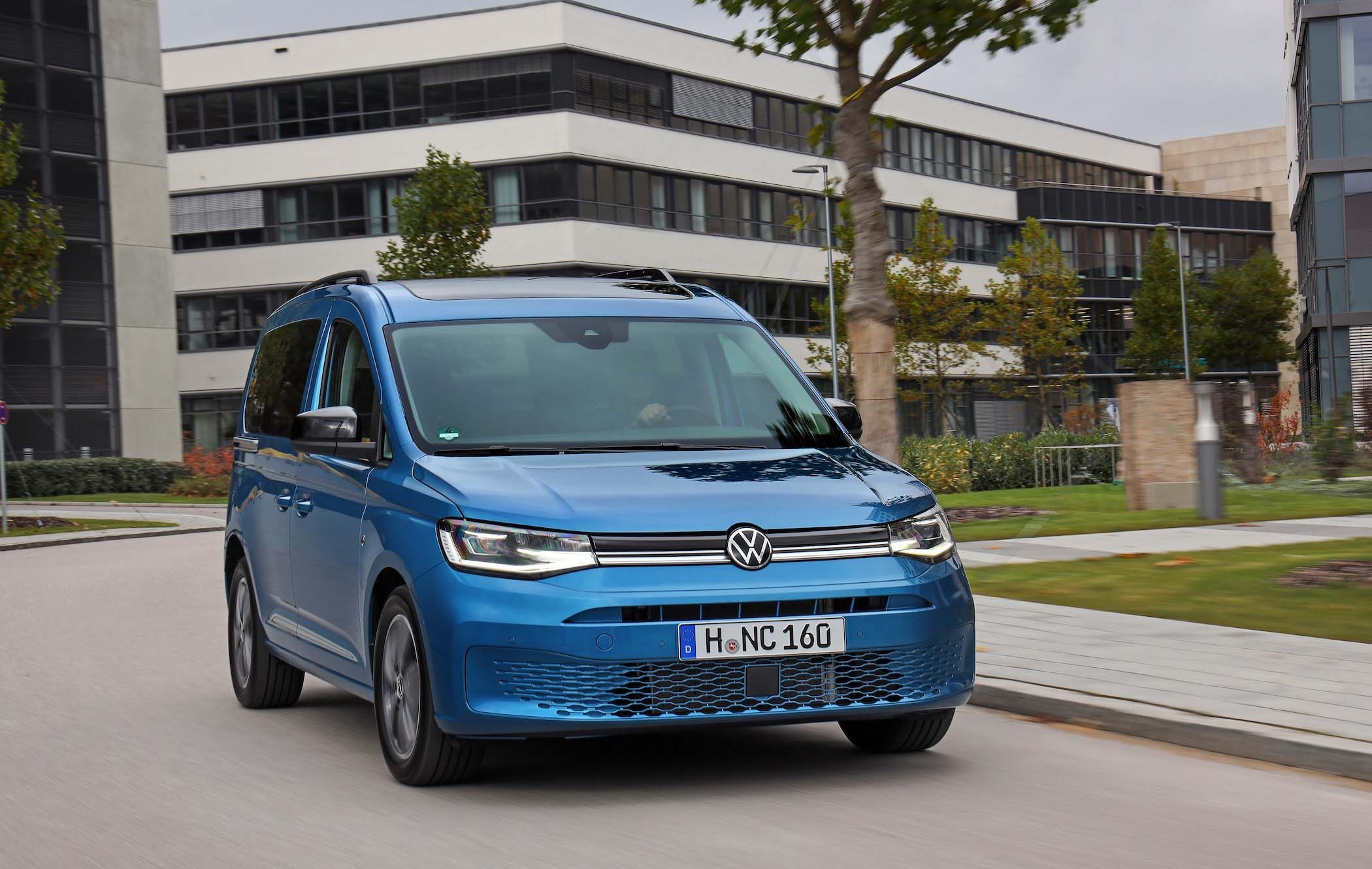Blue Volkswagen Caddy Life driving on a city street with modern buildings and trees in the background.
