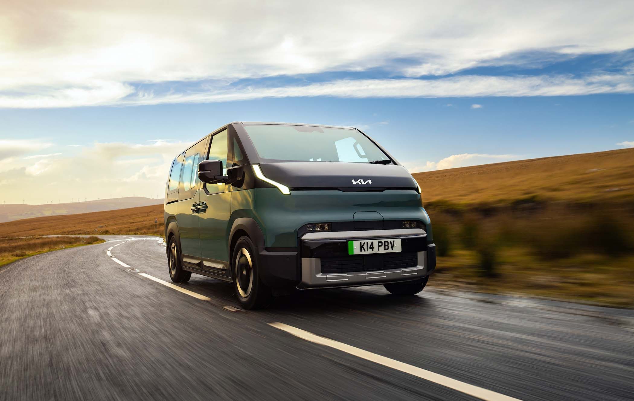 A green Kia PV5 Passenger drives on a wet, winding road through an open, grassy landscape under a partly cloudy sky.