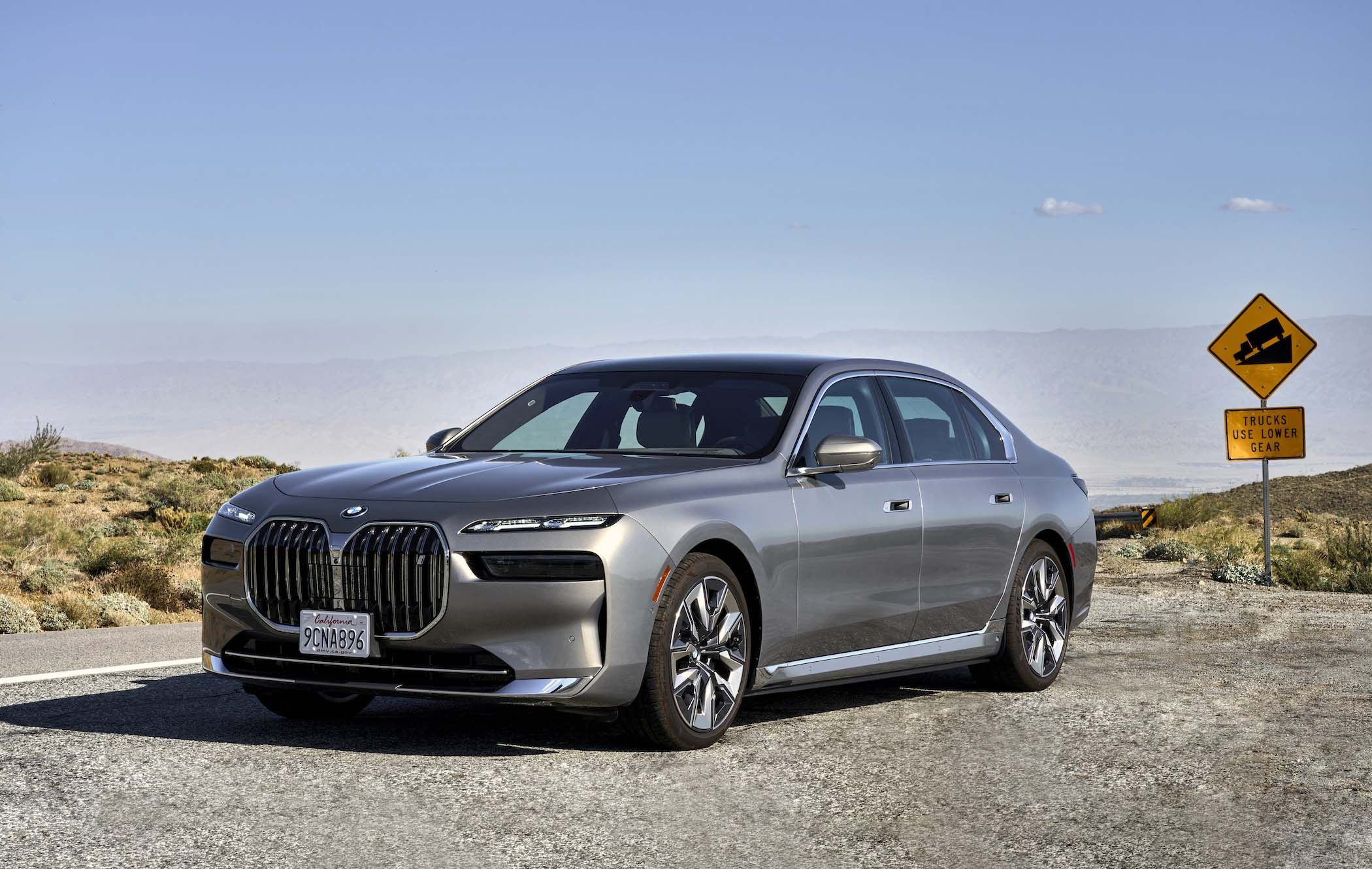 Silver BMW 7 Series parked on a desert road with a "Vehicles in Low Gear" sign and mountains in the background under a clear blue sky.