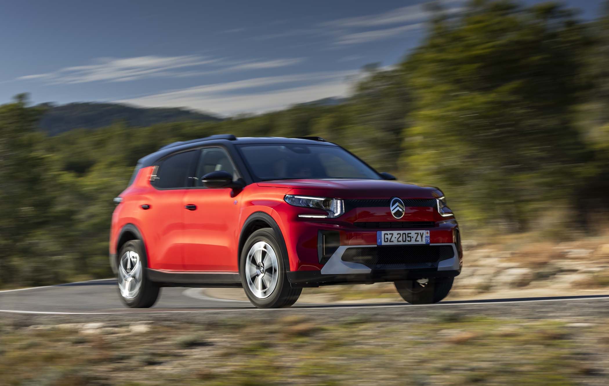 Red Citroën ë-C3 Aircross driving on a curvy road with blurred greenery in the background, under a clear blue sky.