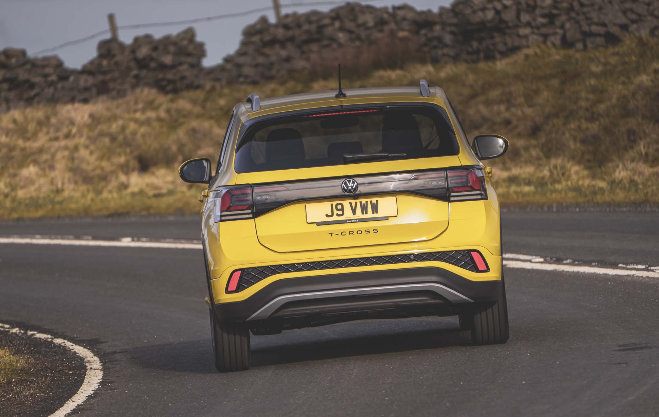 Yellow Volkswagen T-Cross driving on a curved road with a grass and stone wall backdrop.