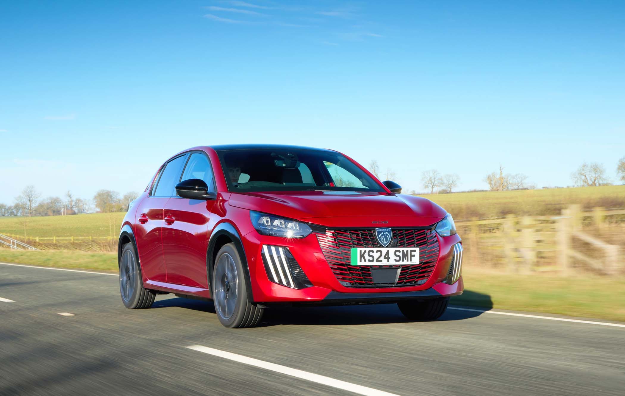 Red Peugeot E-208 driving on a rural road with fields and a clear blue sky in the background.