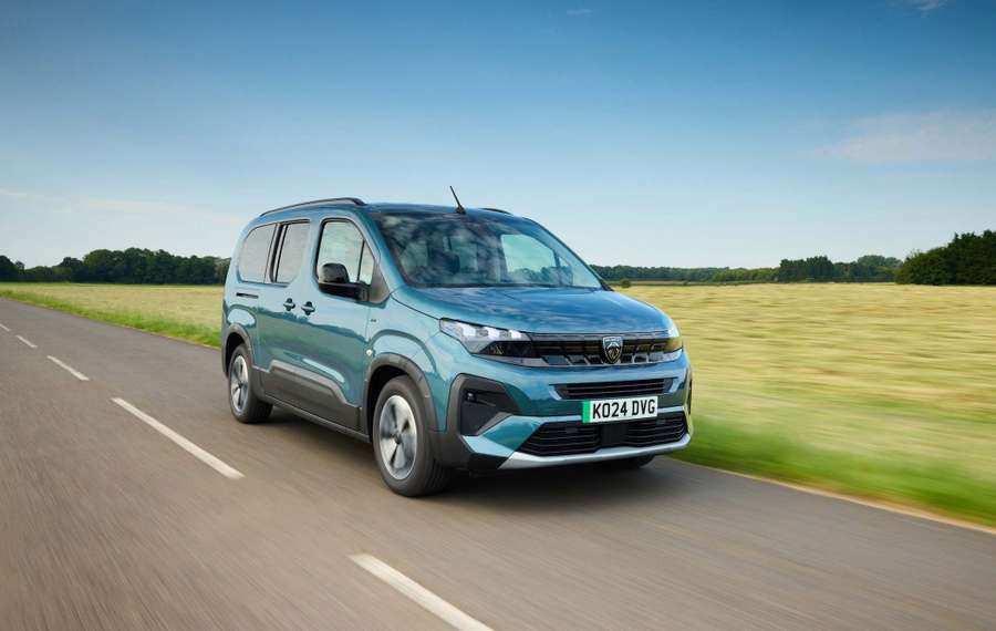 A blue Peugeot e-Rifter drives on a rural road beside a field under a clear blue sky.
