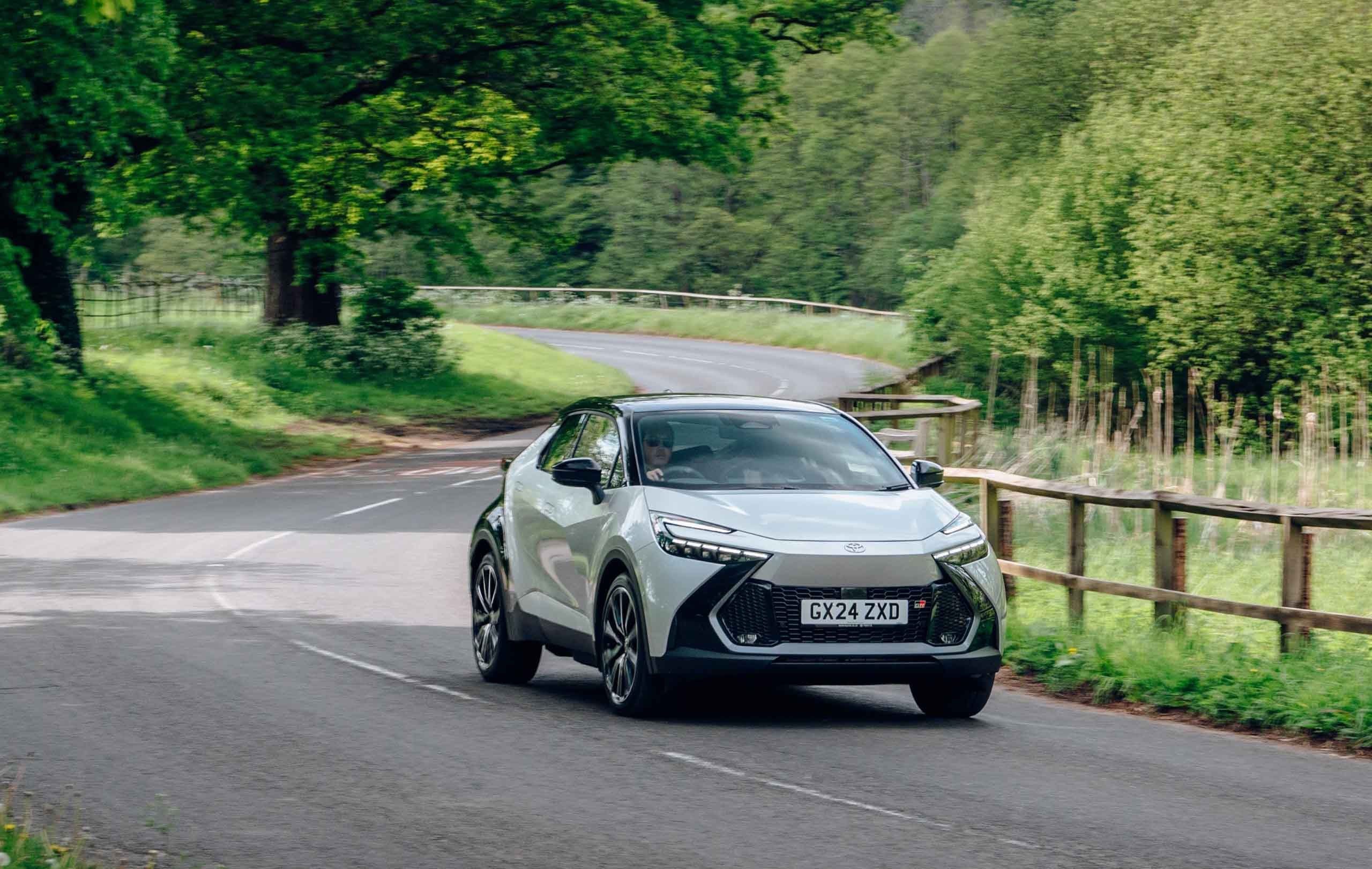 A silver Toyota C-HR driving on a curving country road surrounded by lush green trees and wooden fences.