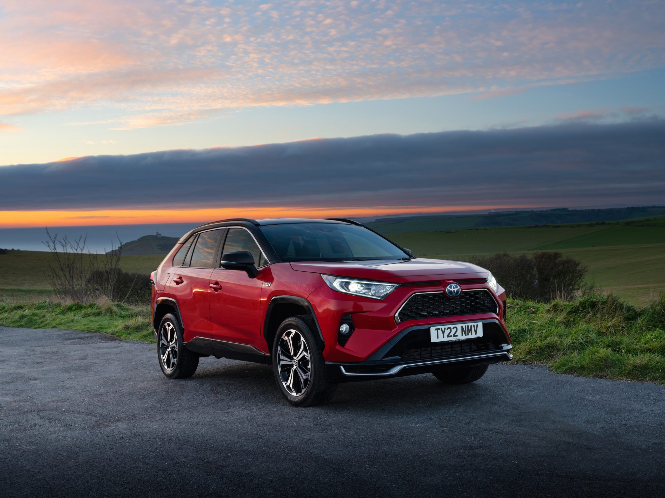 A red Toyota RAV4 hybrid parked on a road next to a sunset