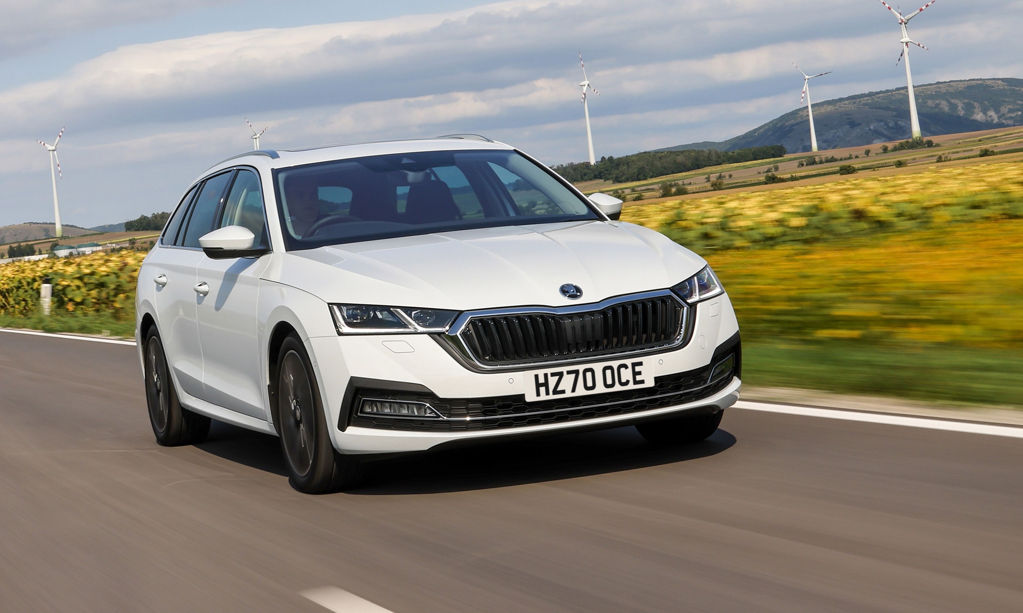 A white Skoda Octavia iV Estate driving on a road next to a wind farm