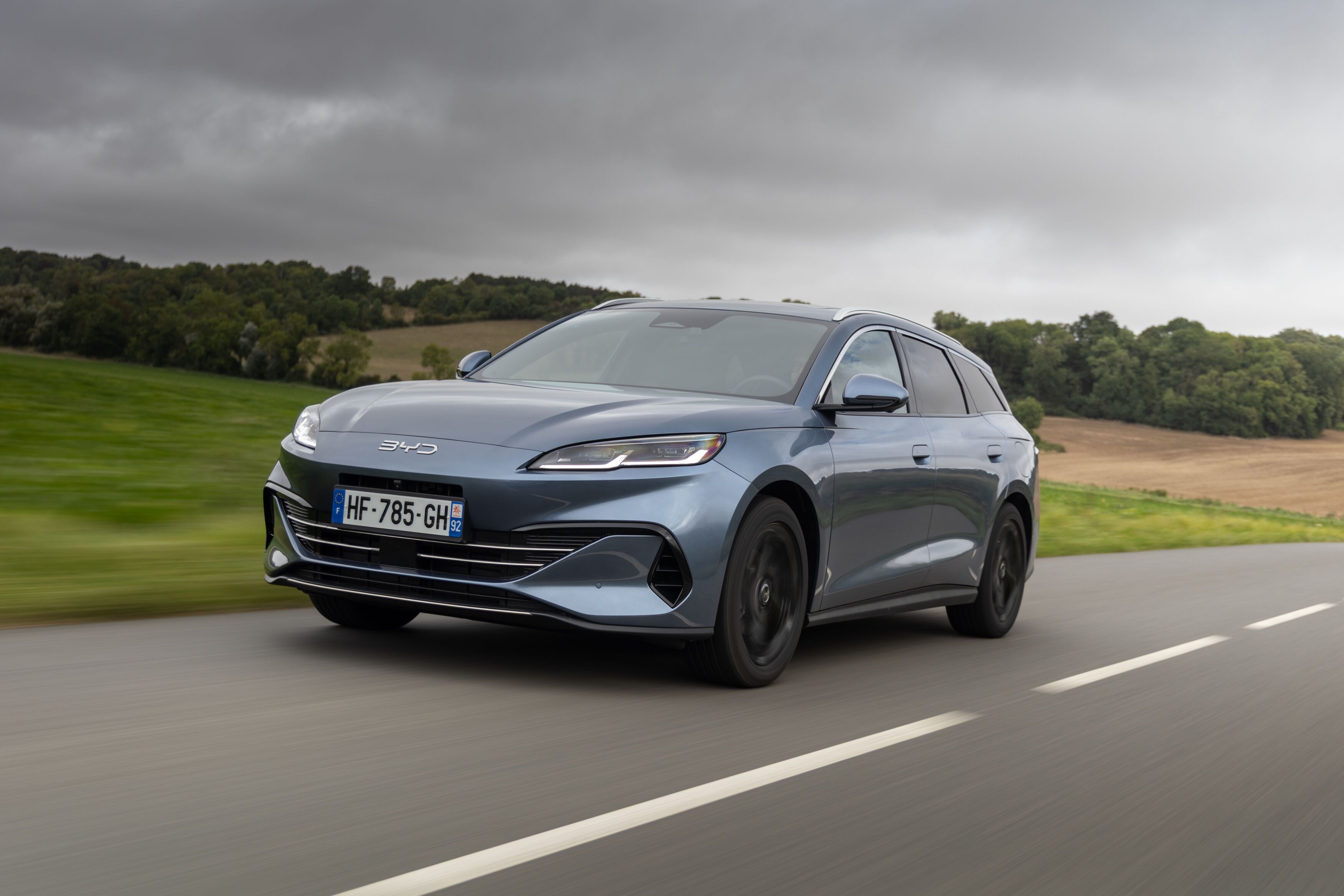 A sleek, silver car drives on a country road under a cloudy sky, with green fields and trees in the background.
