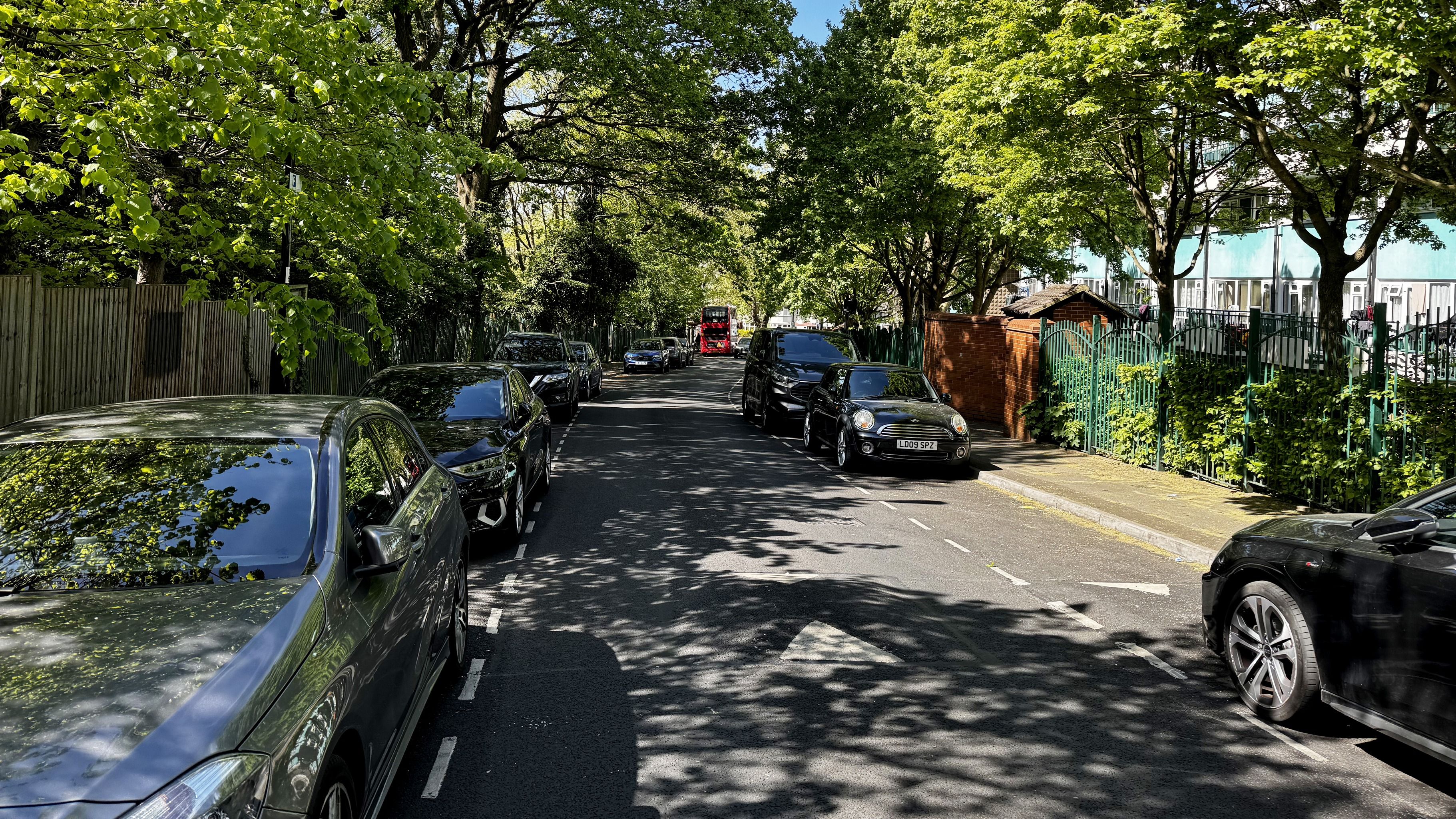 Tree-lined street with parked cars and a red double-decker bus in the distance on a sunny day.