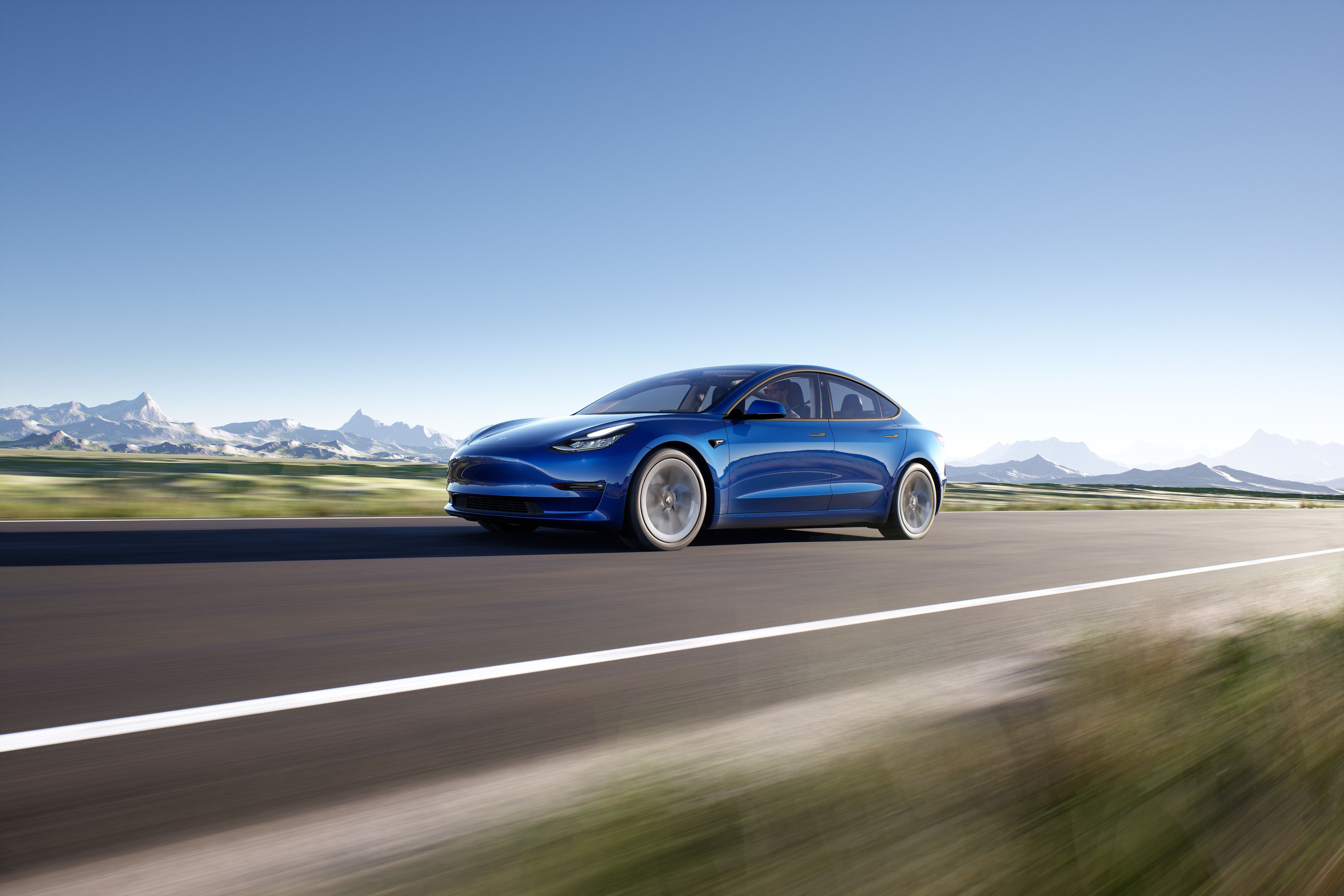 a dark blue tesla model 3 driving on an open road with a snowy mountain range in the distance