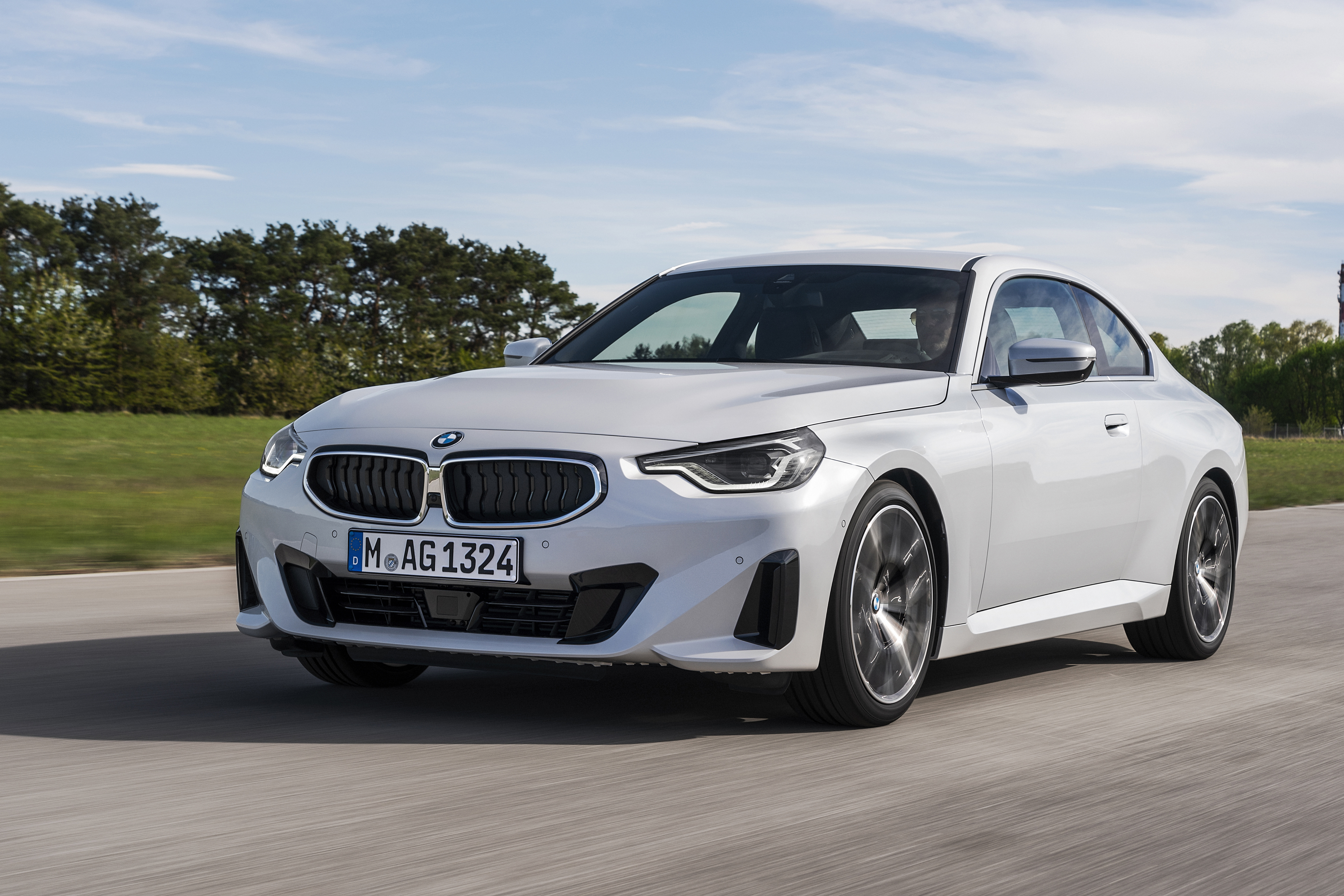 A white BMW 220i driving on a round surrounded by trees