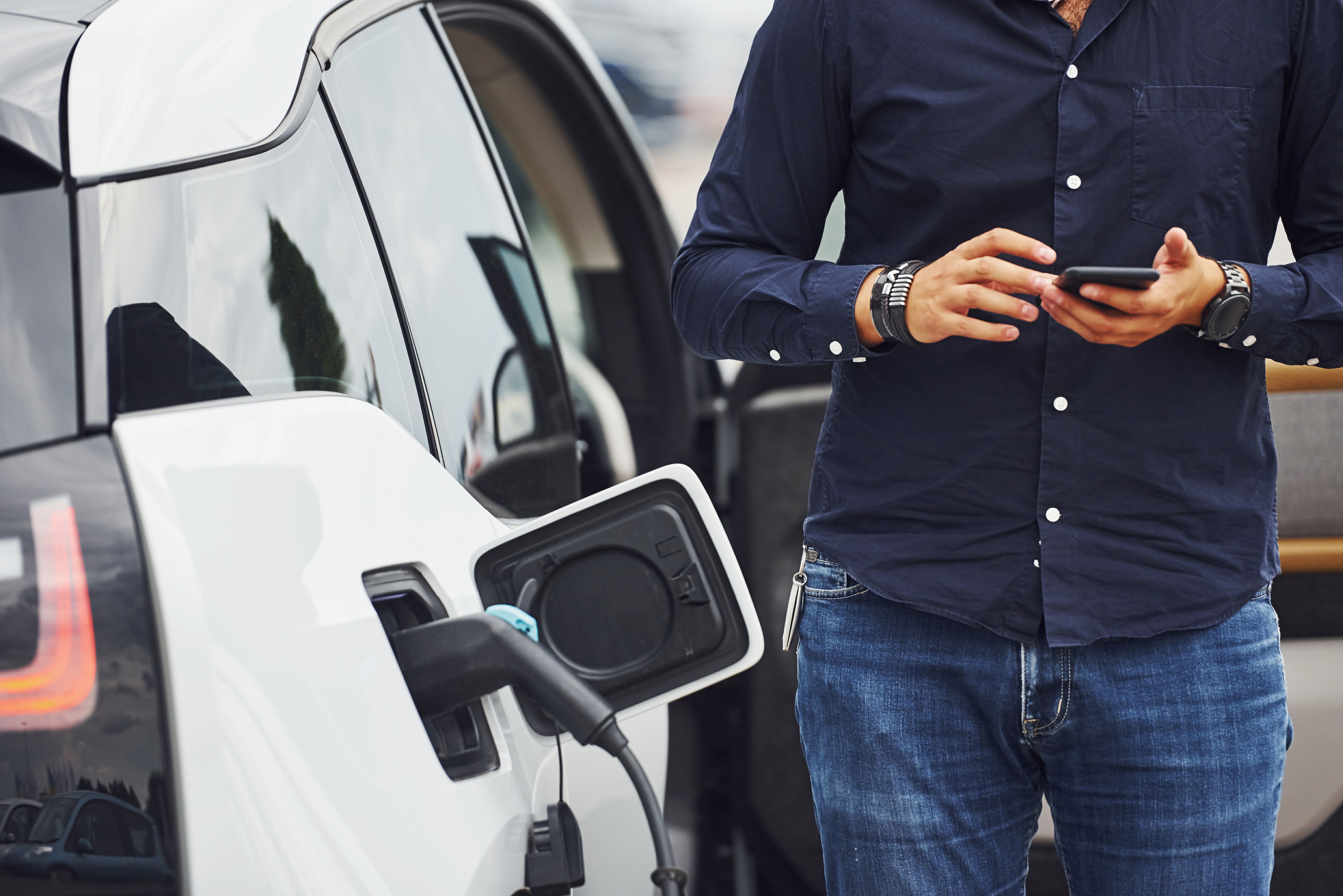 A close up of a charger plugged into an EV and the body of a man using his phone