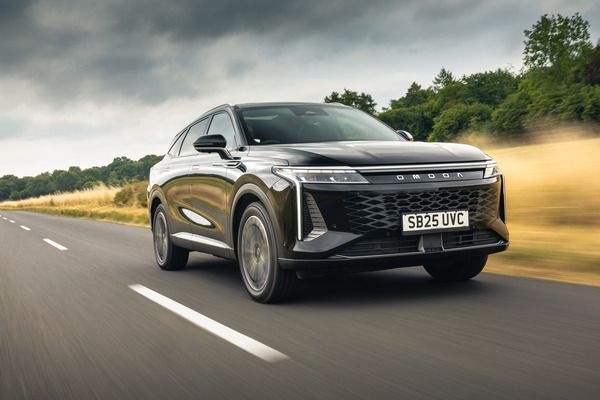 Black OMODA 9 SUV driving on a rural road with grassy fields and trees under a cloudy sky.