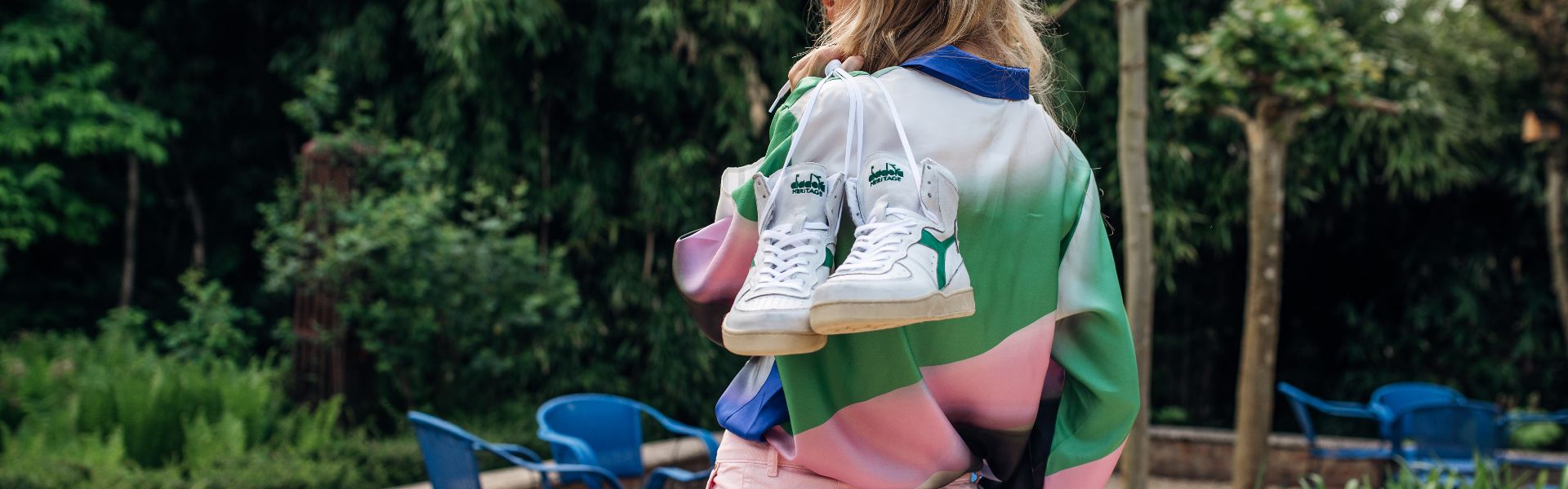 Vrouw in sportieve kleding met rug naar het beeld gekeerd maar met een paar Diadora Heritage sneakers over haar schouder bungelend