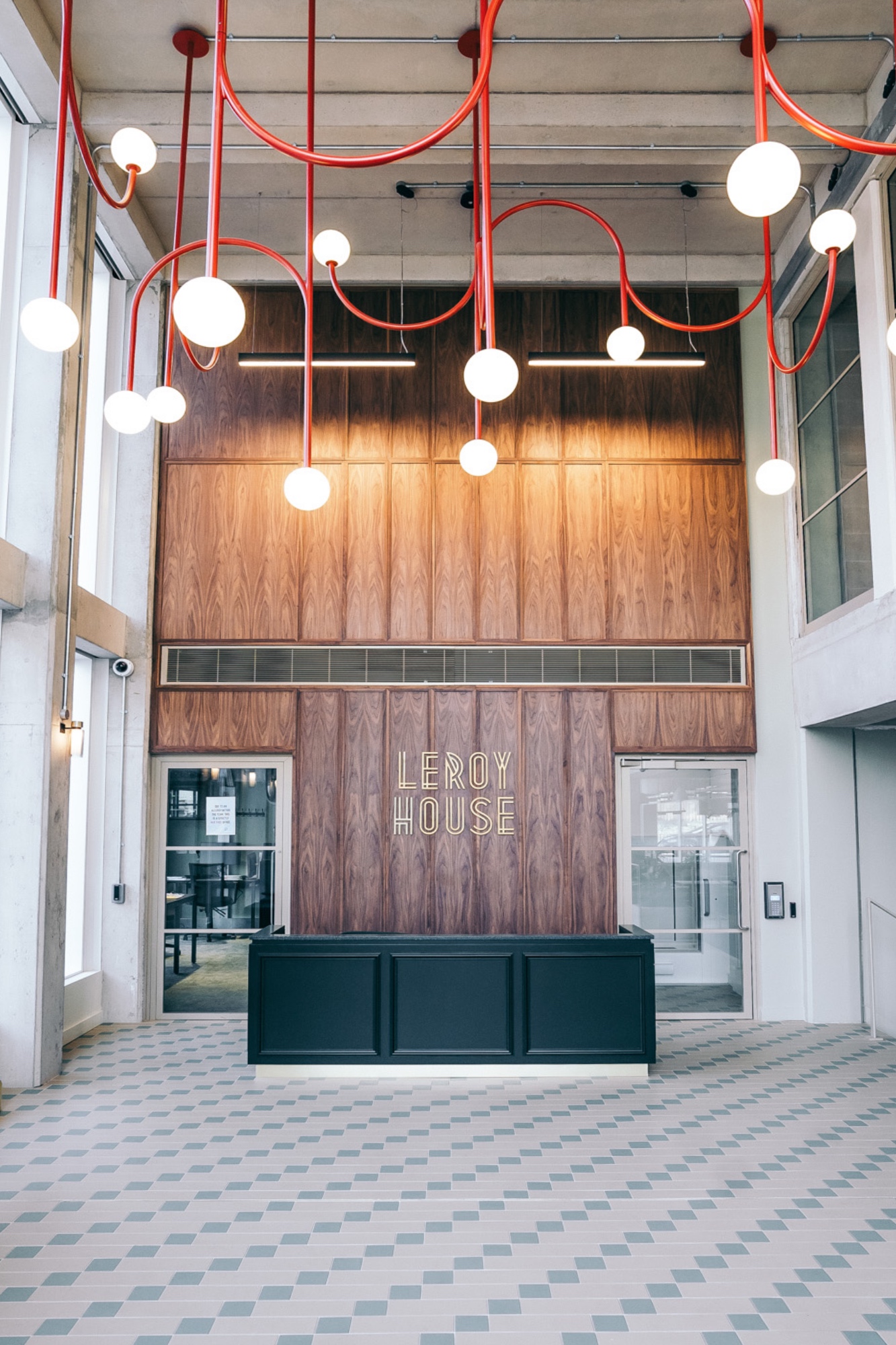 Modern lobby at Leroy House with geometric floor tiles, a dark reception desk, and red ceiling lights. "Leroy House" is displayed in art deco style on the wooden wall.