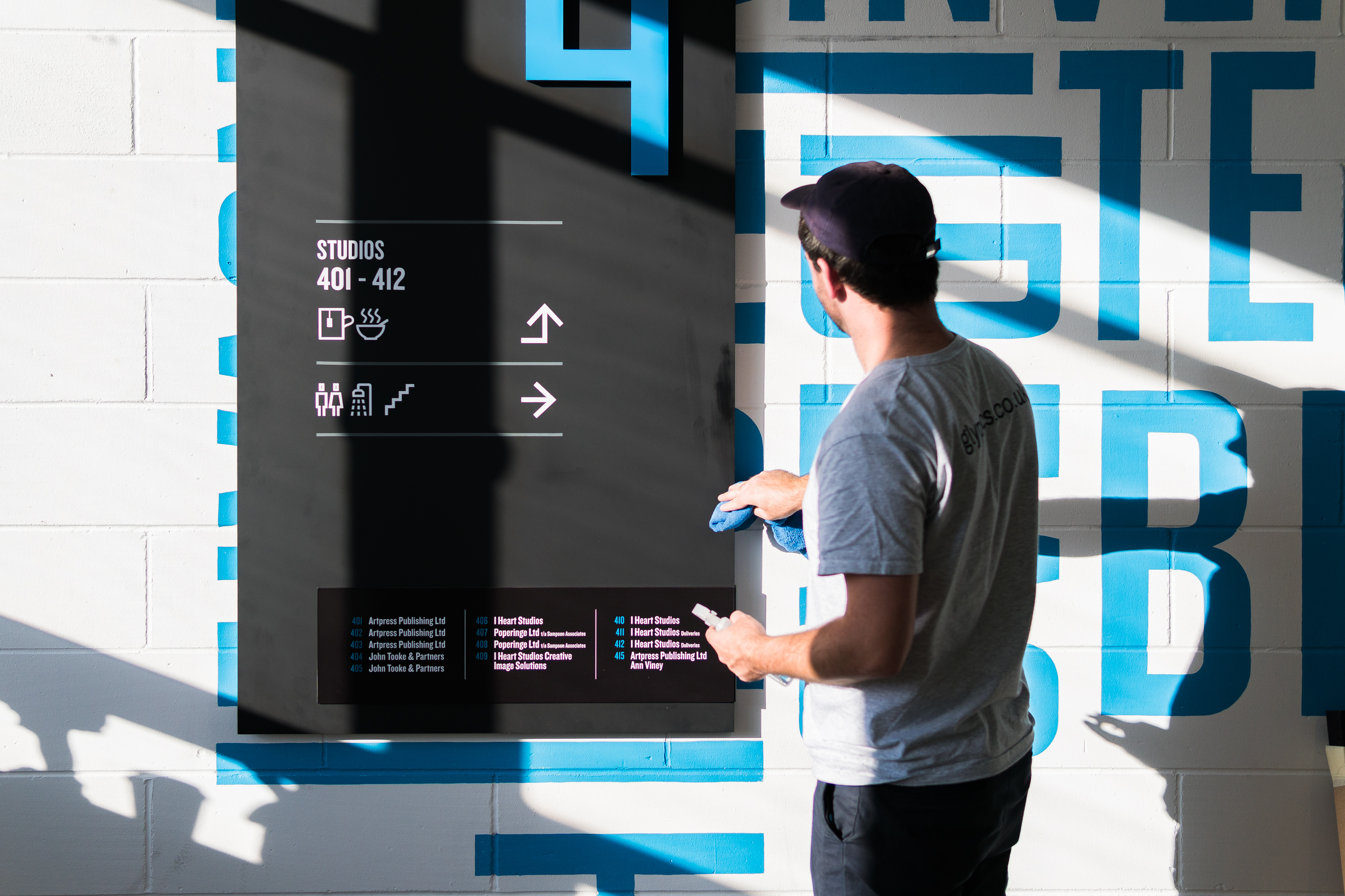 Glyphics sign installer using a blue cloth to tidy a large black directory sign on a white wall with blue lettering and shadows, at the Biscuit Factory co-working space..
