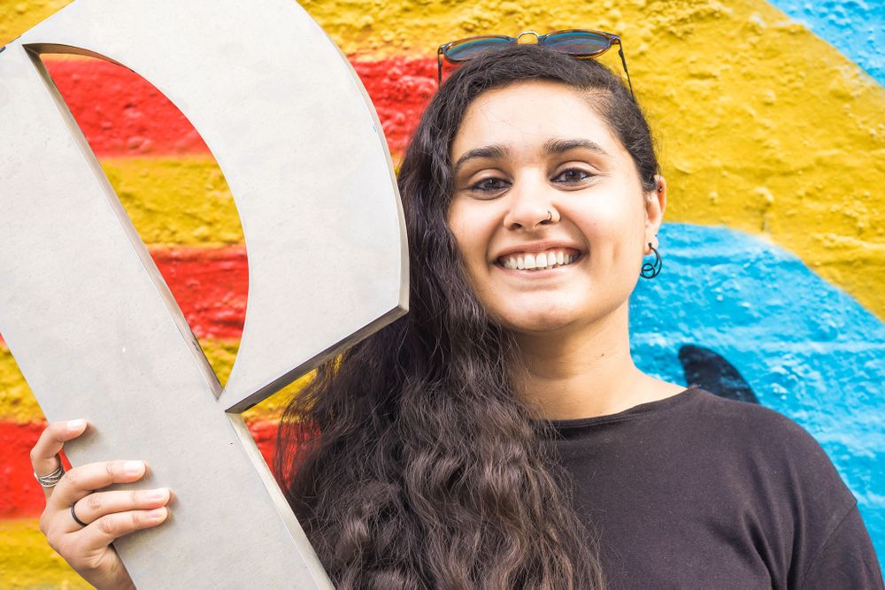 Amrita holding a large silver P in front of colourful street art in Shoreditch