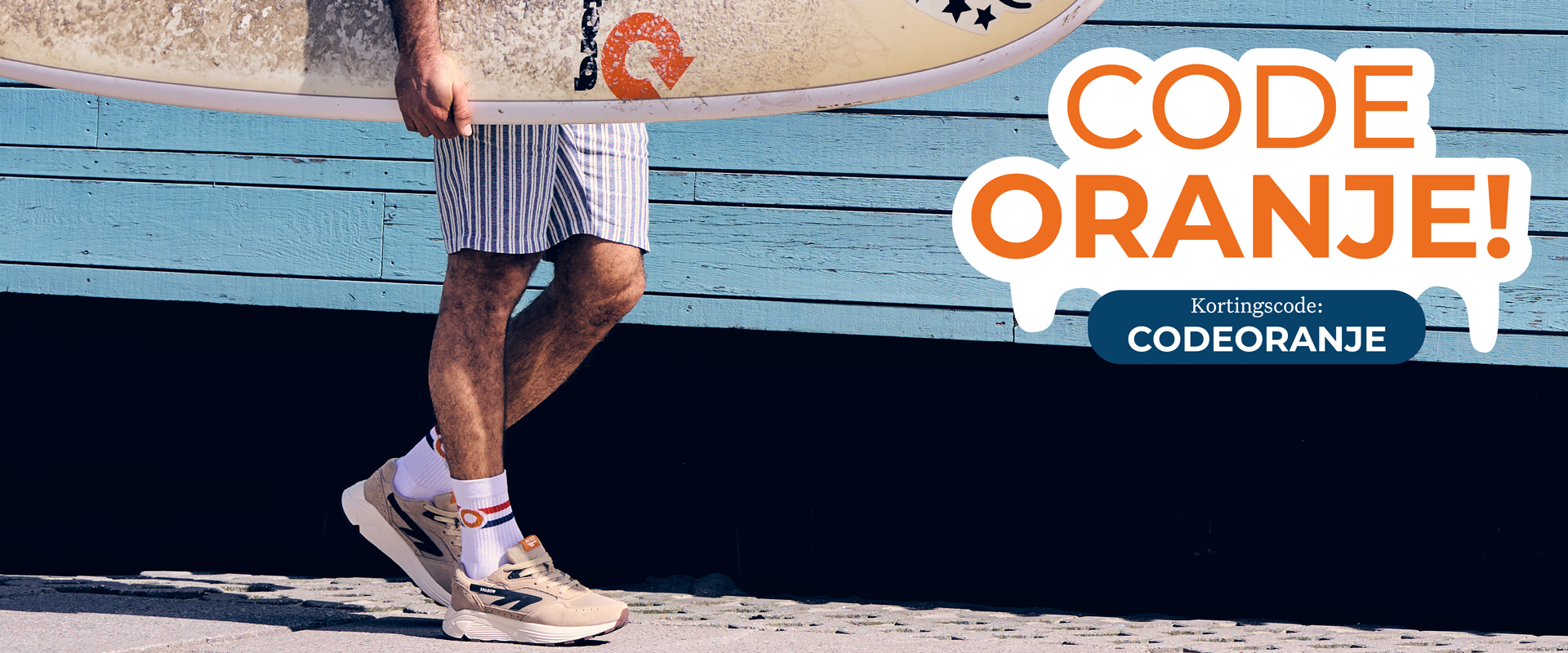 Man in striped shorts and sneakers holding a surfboard walks by a blue wall. Text reads "Code Oranje! Kortingscode: CODEORANJE".
