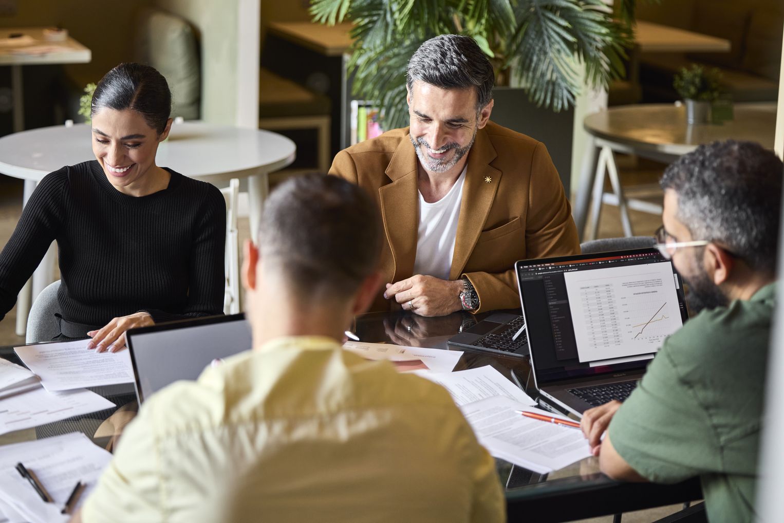 Vier Personen sitzen an einem Glastisch in einem hellen, modernen Büro/Café. Auf dem Tisch: zwei aufgeklappte Laptops (einer zeigt eine Tabelle mit Diagramm), Papierdokumente, Stifte. Die Gruppe wirkt kollaborativ, entspannt und fokussiert – ein Mann in beigem Sakko und eine Frau in Schwarz lächeln, im Vordergrund zwei weitere Teammitglieder von hinten/seitlich.