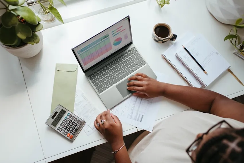 Person using a laptop with charts on the screen, surrounded by a notebook, calculator, coffee cup, and plants on a white desk.