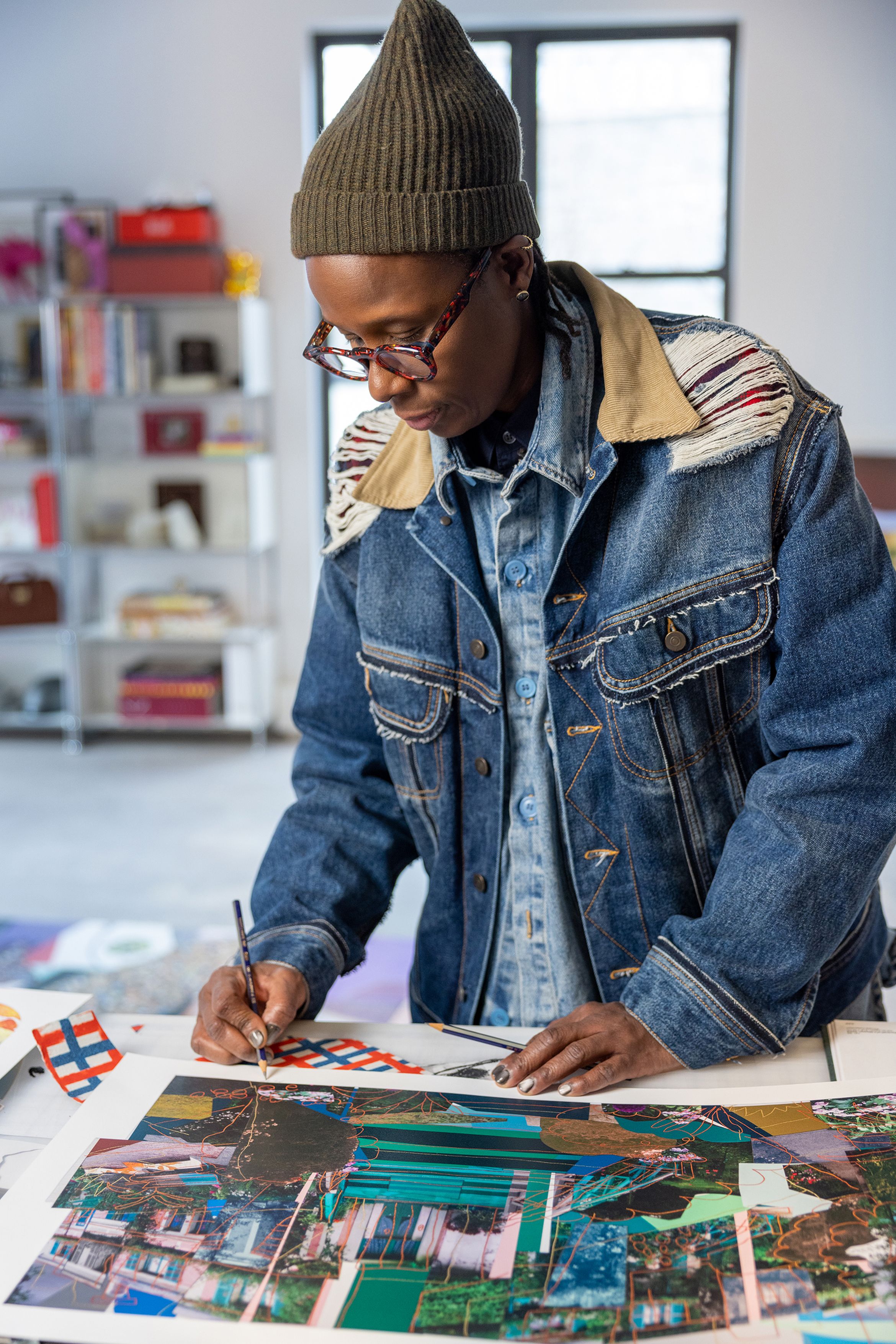 Person wearing a denim jacket and beanie, focused on creating artwork at a table in a creative studio.
