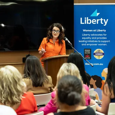 A woman in an orange blouse speaks at a podium in front of an audience. A banner for "Women at Liberty" is displayed beside her.