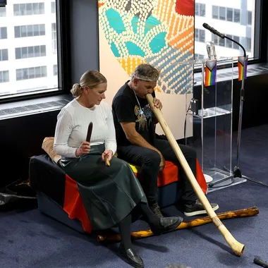 Two people perform with traditional instruments indoors, seated on a bench with a colorful backdrop, and a microphone stand nearby.