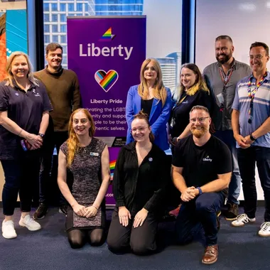 A group of people posing in front of a "Liberty Pride" banner and a rainbow flag, smiling in a bright room.