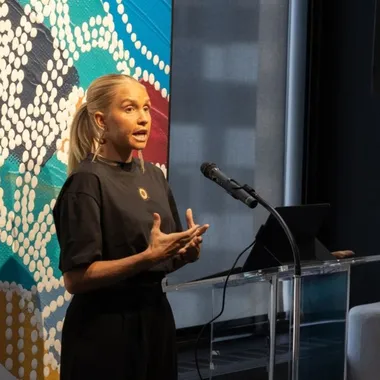 A woman speaks at a podium with a microphone, gesturing with her hands. A colorful abstract painting is in the background.