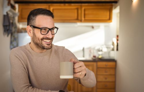 A man with glasses in a beige sweater is sitting in a bright kitchen, holding a mug with one hand. The background shows wooden cabinets and sunlight coming in through a window.