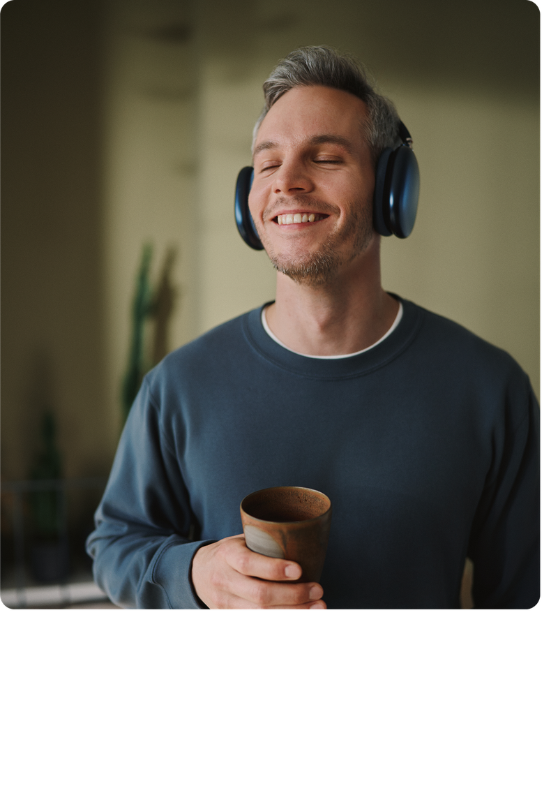 Smiling man with headphones holds a mug, standing indoors in a relaxed setting.