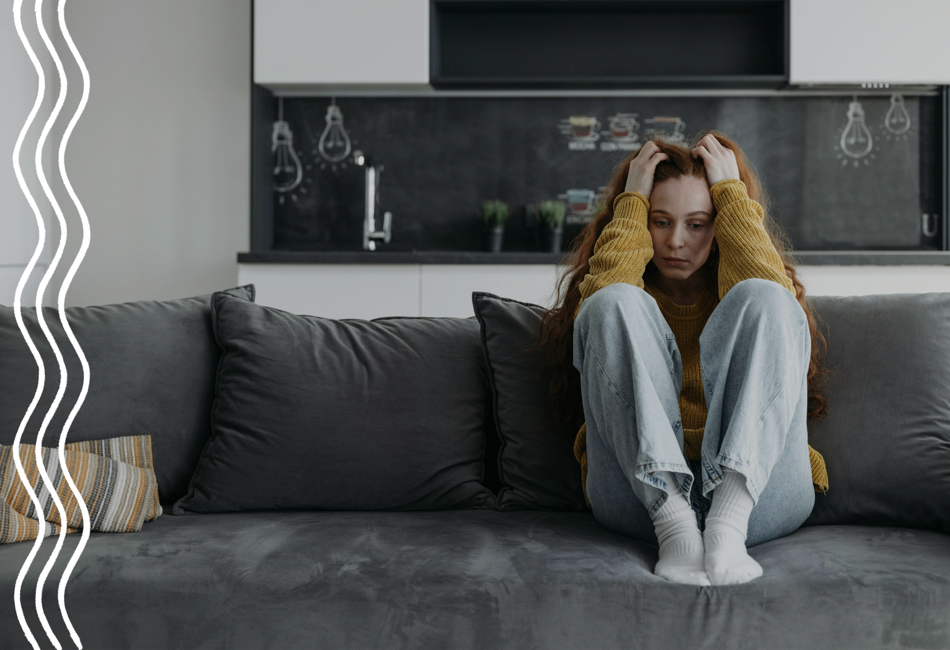 Eine Frau mit roten Haaren, gelben Pullover und heller Jeans sitzt auch einem grauen Sofa, schaut mit ernster Miene nach unten und fährt mit ihren Händen durch ihre Haare.