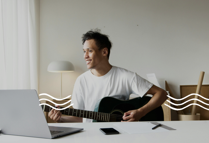 Ein Mann im weißen T-Shirt sitzt mit einer schwarzen Gitarre am Schreibtisch, auf dem weißes Papier, ein Handy und ein aufgeklappter Laptop steht.