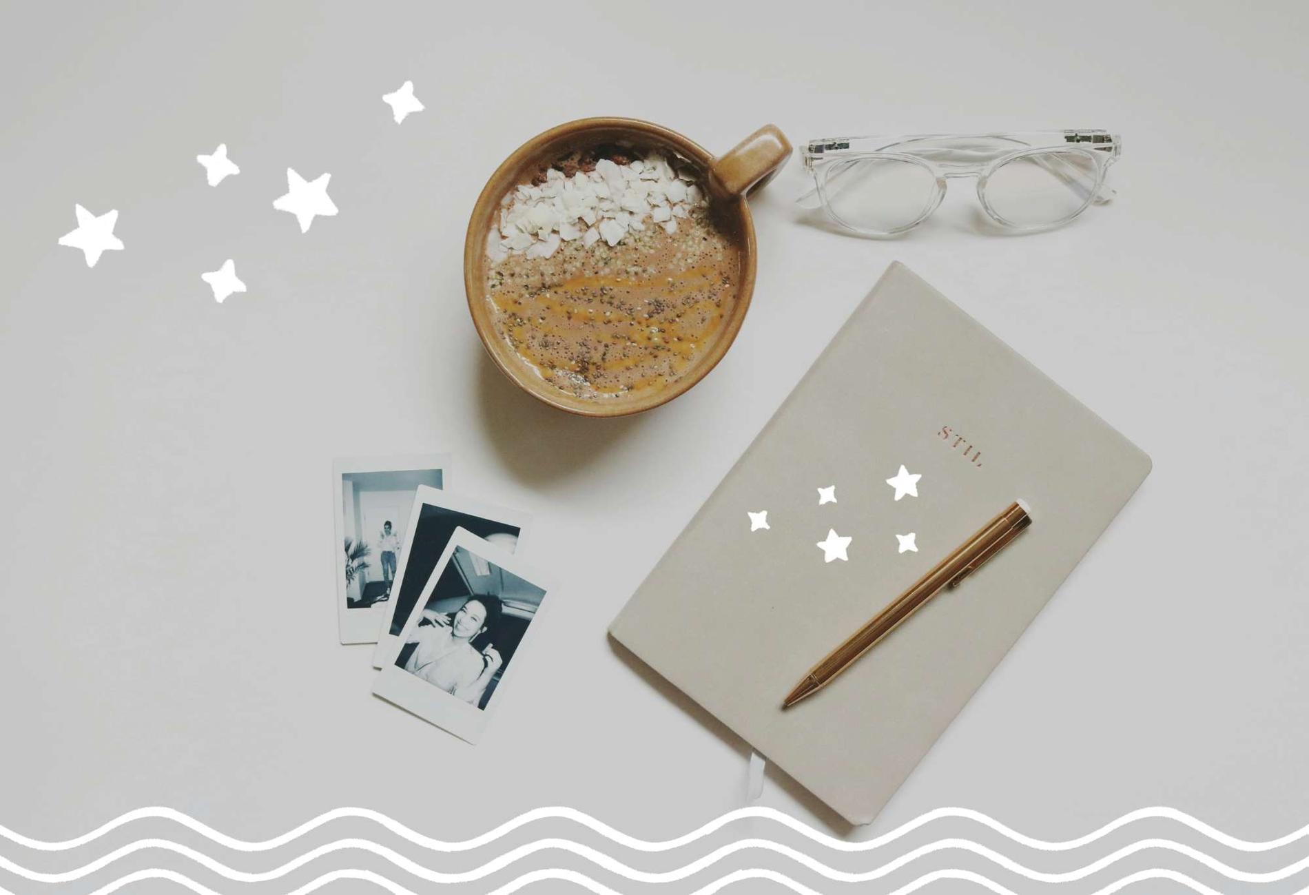 Auf einem weißen Hintergrund liegen ein paar Polaroid-Fotos, eine Tasse heiße Schokolade, eine Brille und ein Journal mit Stift.