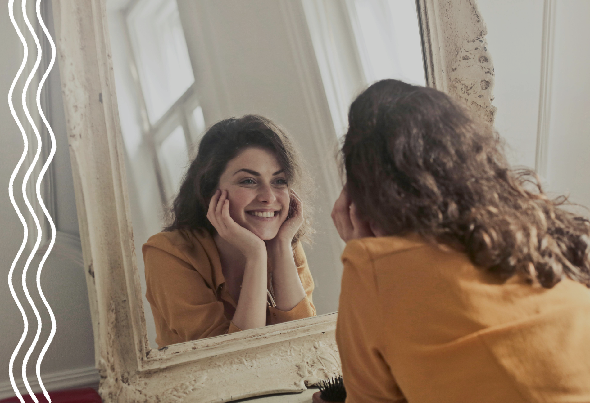Eine Frau mit braunen Locken und einer gelben Bluse schaut sich in einem Spiegel an, stützt ihr Gesicht auf ihren Händen ab und lacht.
