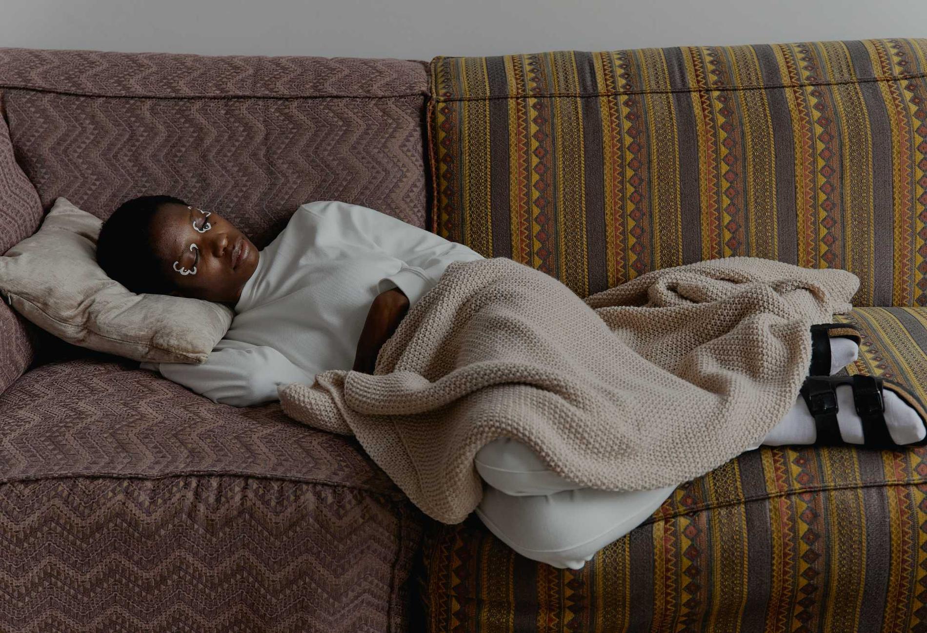 Eine Schwarze Person liegt mit einer beigen Decke auf einem gemusterten Sofa und schläft ruhig.