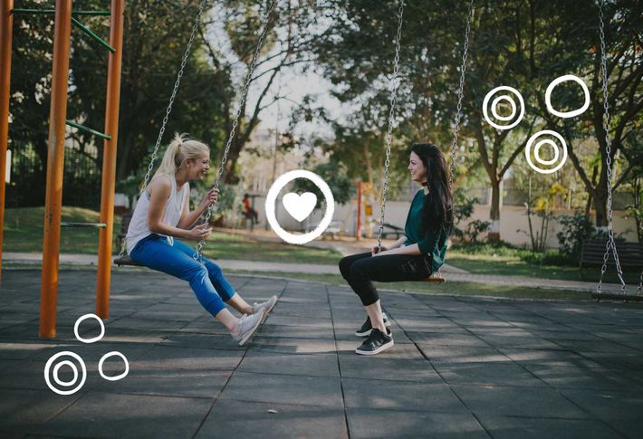 Zwei junge Frauen sitzen sich seitlich auf jeweils einer Schaukel gegenüber und lachen sich an. Im Hintergrund ist ein grüner Park zu sehen.
