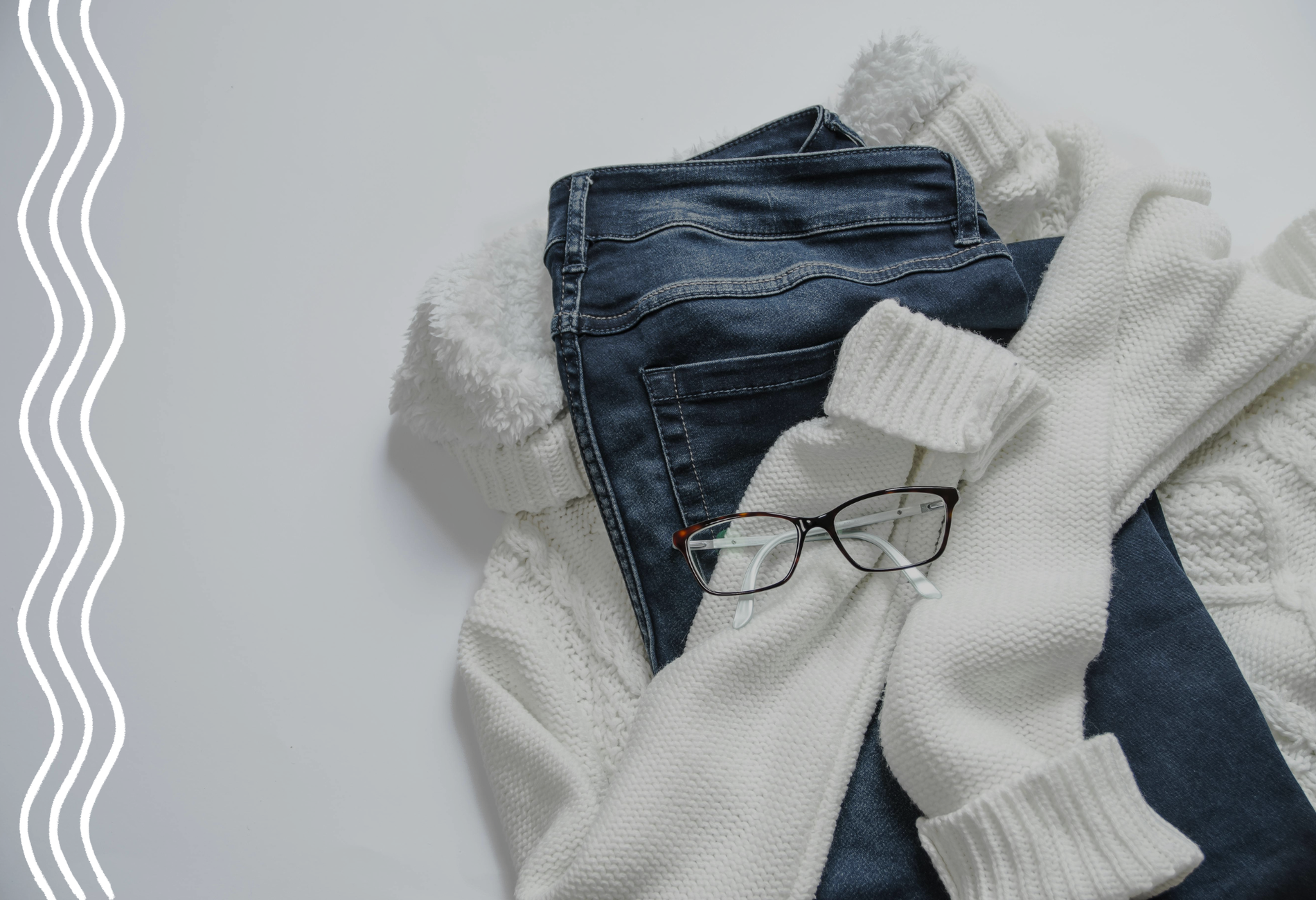 Auf einem weißen Untergrund liegen ein weißer Rollkragen-Strickpullover, eine dunkle Jeanshose zusammengefaltet und ganz oben eine Brille.
