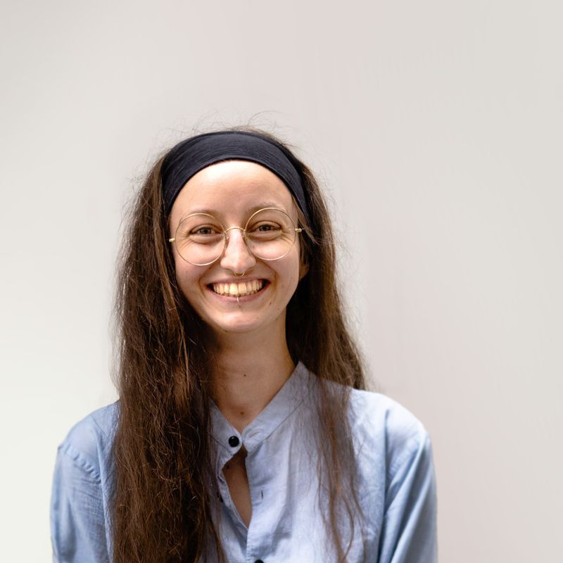Eine junge Frau mit langen braunen Haaren und schwarzem Stirnband trägt eine runde Brille und lächelt strahlend. Sie hat ein hellblaues Hemd an und steht vor einem neutralen hellen Hintergrund.