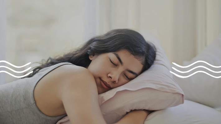 Eine Frau schläft zufrieden in ihrem hellen Zimmer. Sie hat schwarze Haare, liegt seitlich auf dem Bauch, umarmt ein Kopfkissen und trägt ein graues Top.