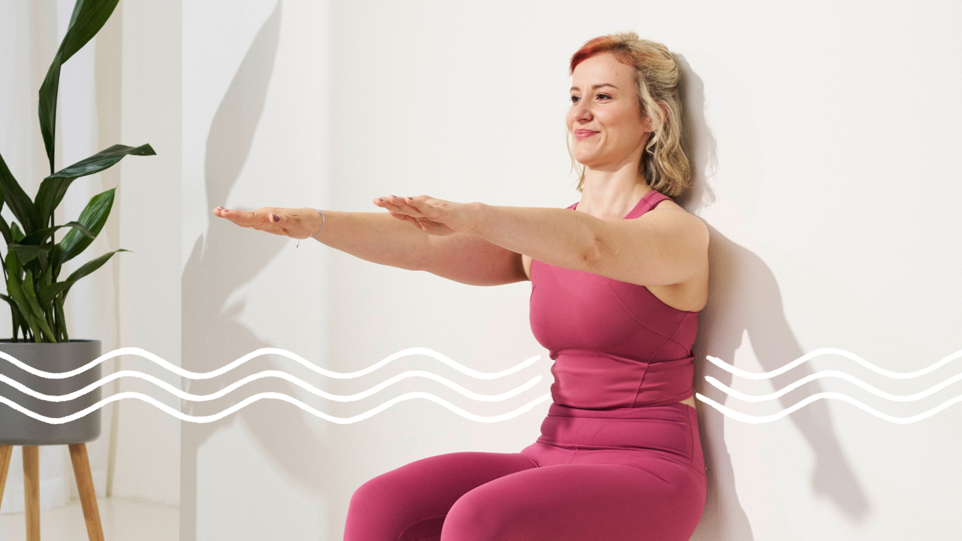 Eine Frau mit blonden, schulterlangen Haaren in einem pinken Pilates-Outfit sitzt im Wall Squat, mit dem Rücken an die Wand gelehnt und den Beinen im rechten Winkel. Die Arme sind ausgestreckt. Links im Hintergrund ist eine grüne Pflanze.