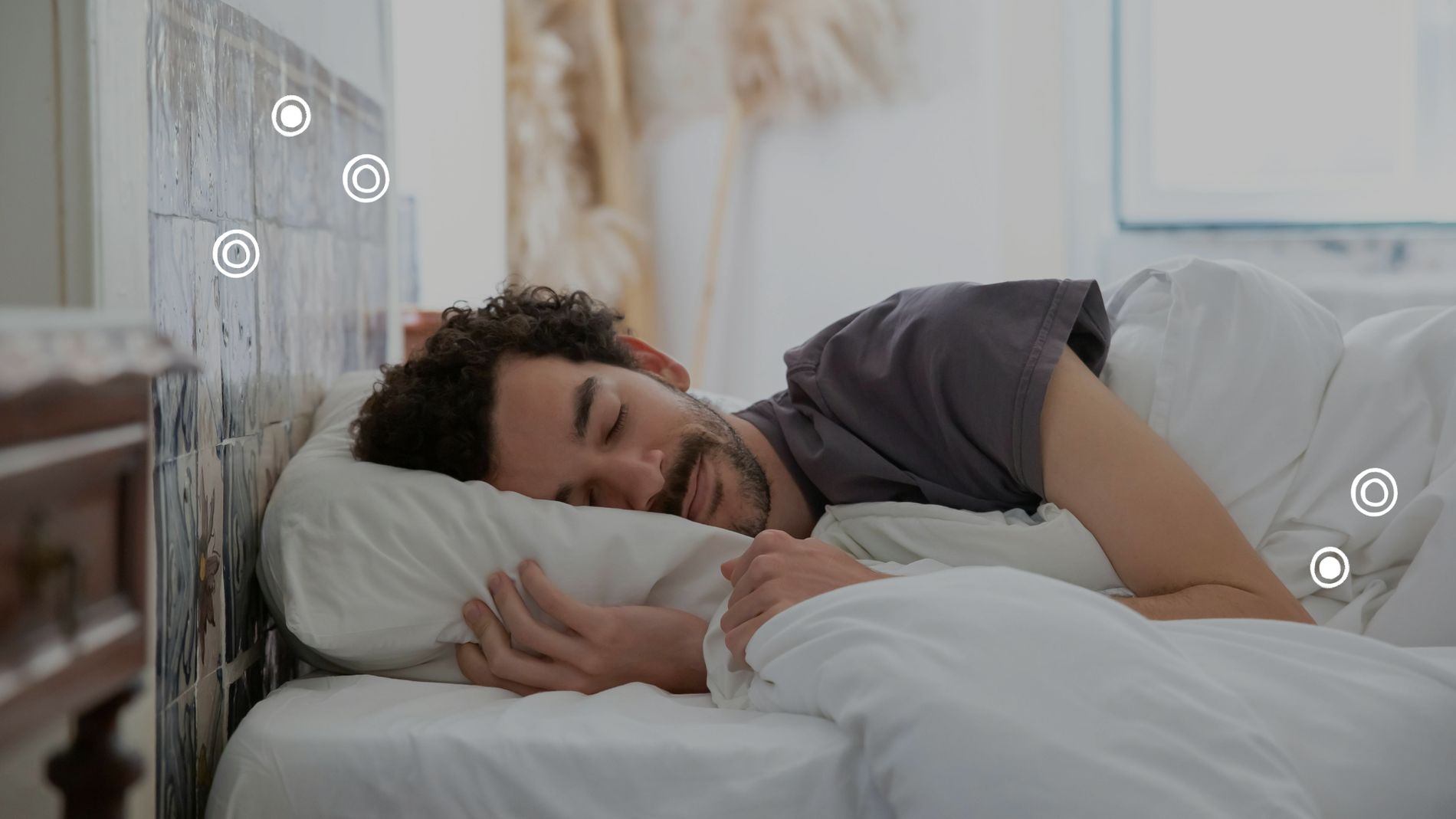 Ein Mann mit dunkelbraunen locken und Bart im grauen T-Shirt liegt in einem Bett mit weißer Bettwäsche auf seiner rechten Seite und schläft.