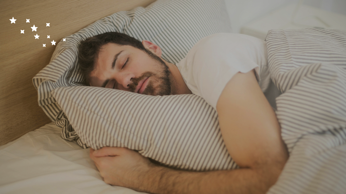 Ein Mann im weißen T-Shirt liegt auf der Seite im Bett mit gestreifter Bettwäsche und schläft.